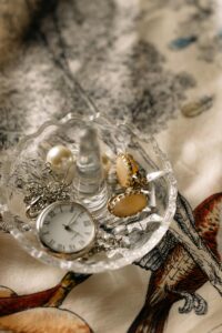 A beautiful display of jewelry and a pocket watch in a crystal dish on a patterned fabric.